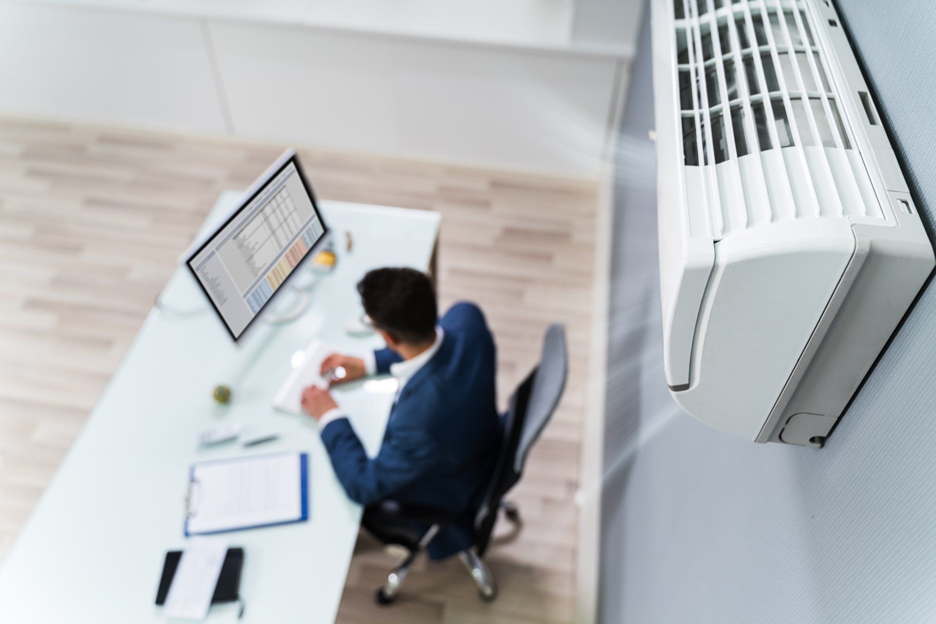 Person in a suit working at a desk with a computer, near a wall-mounted air conditioner.