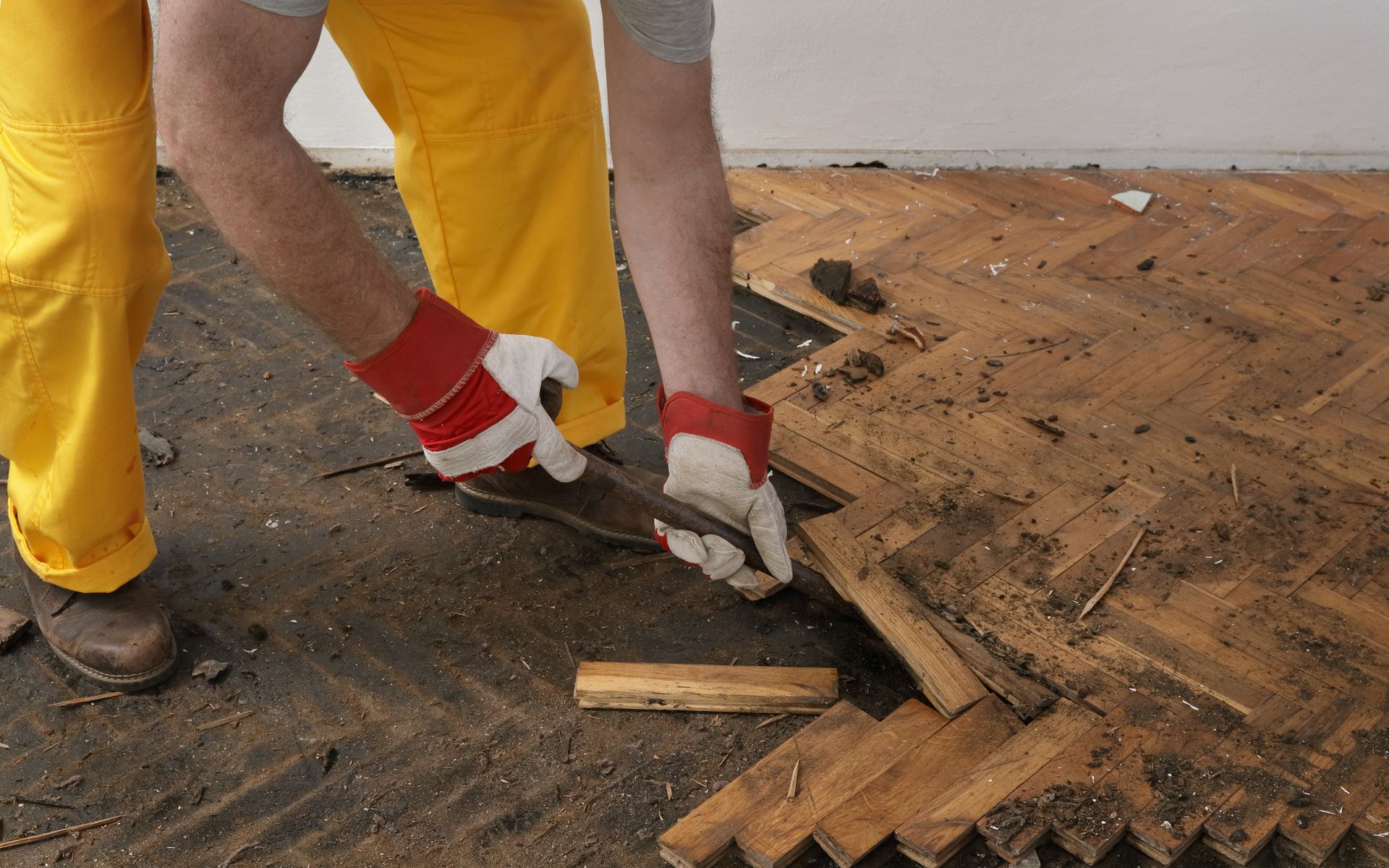 Worker in yellow pants prying up damaged floorboards with a crowbar and gloves