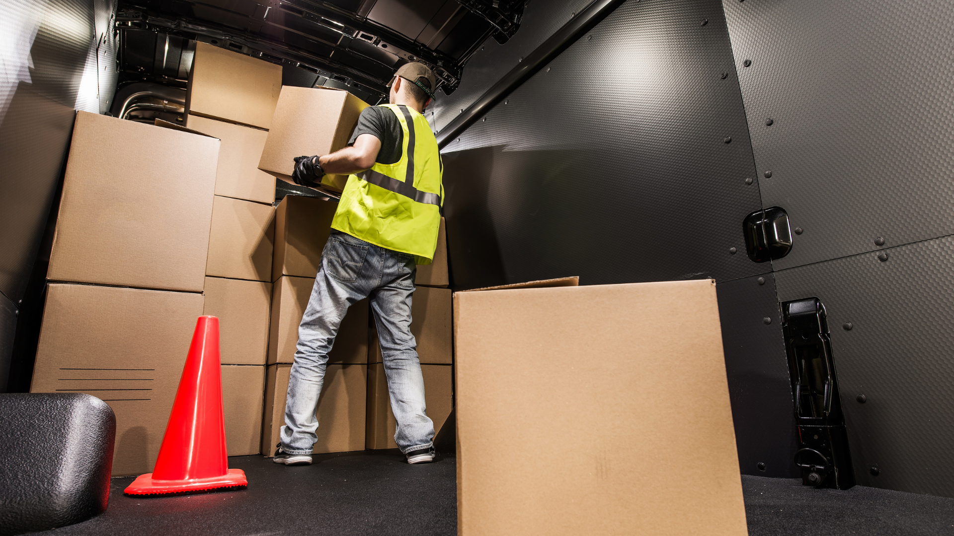 Delivery person in a cargo van wearing a safety vest, handling cardboard boxes. A traffic cone sits nearby.