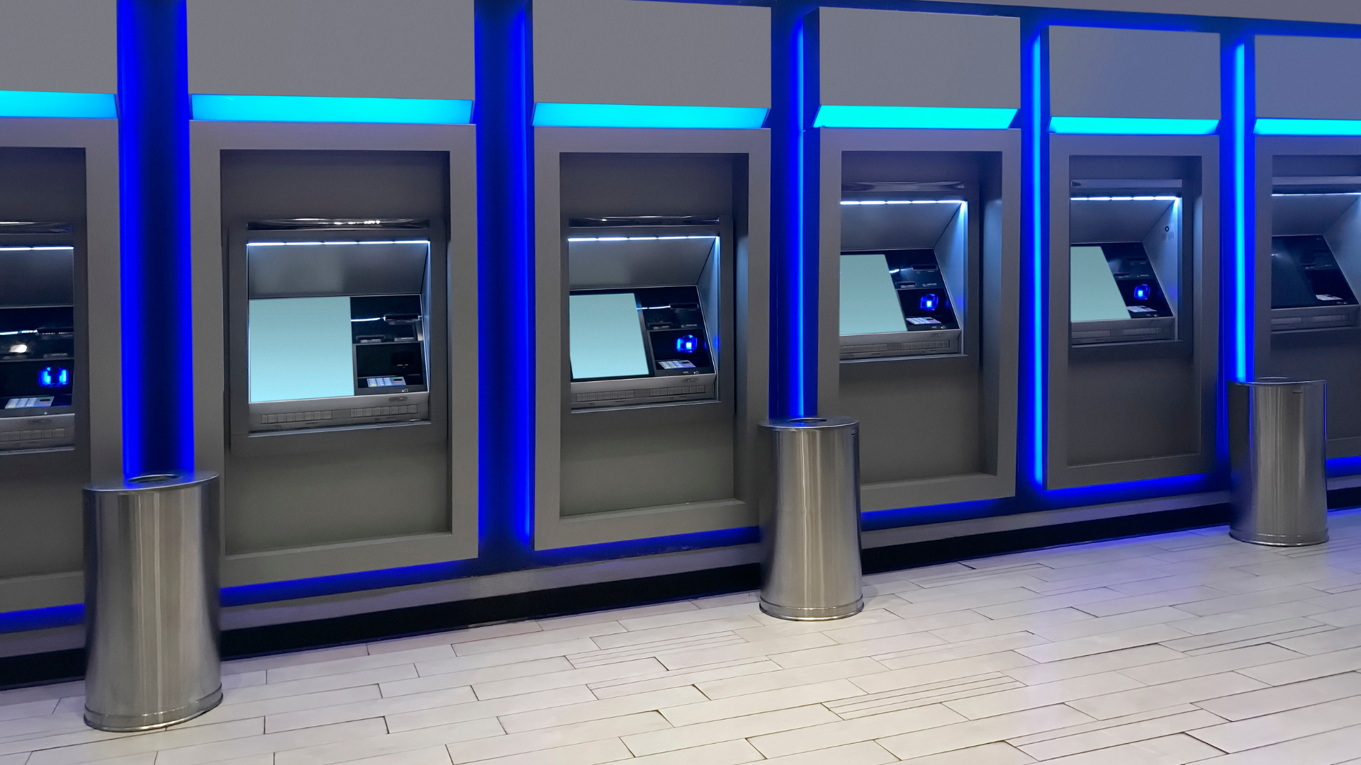 Row of illuminated ATMs in a public space, with blue lighting accents and stainless steel supports.