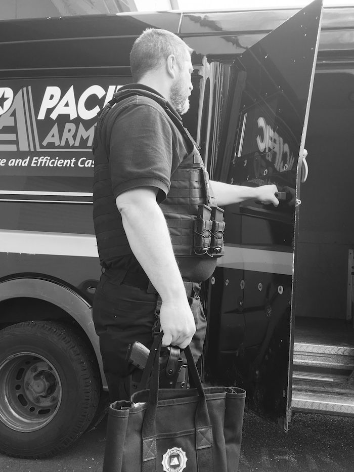 Man in tactical vest opening armored van door, holding a bag and a handgun. Pacific Armor logo visible on the van.