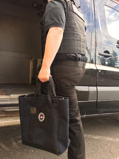 Person in a bulletproof vest and black uniform carries a black tote bag near a black van.