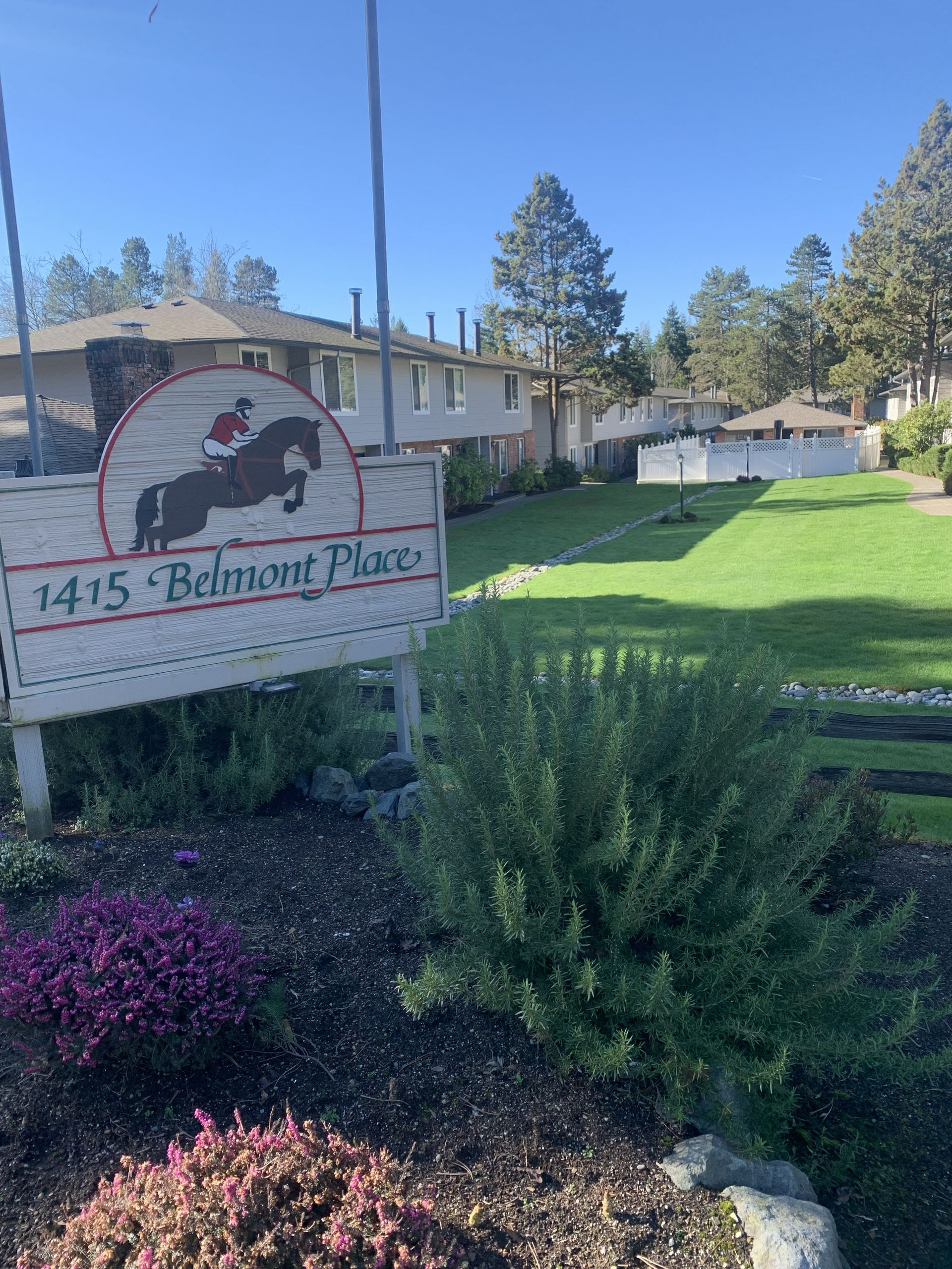 Sign for 1415 Belmont Place apartments, with horse and rider logo. Green lawn and buildings in background. Sunny day.