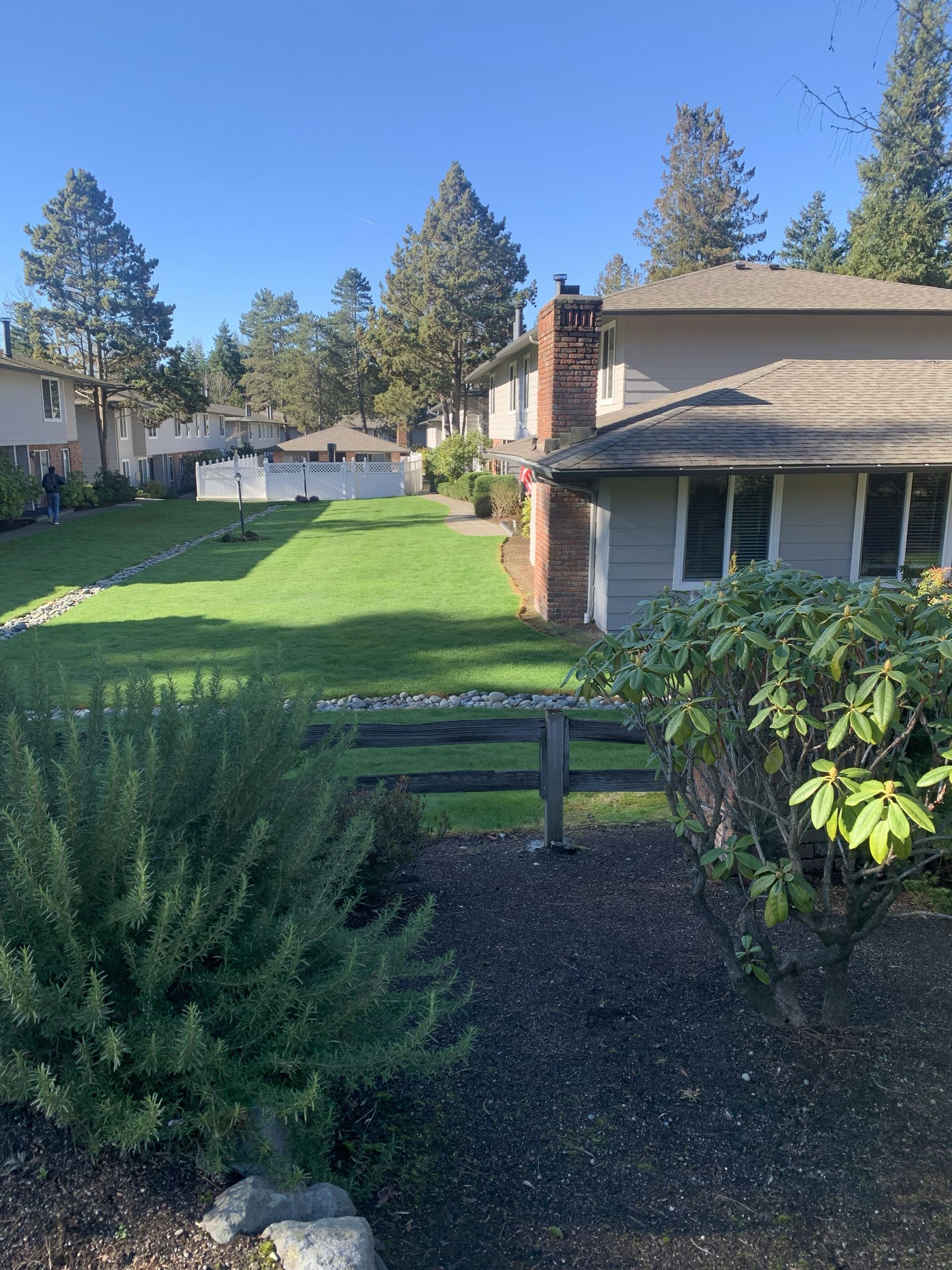 Residential neighborhood with green lawns, trees, and buildings on a sunny day. A wooden fence is in the foreground.