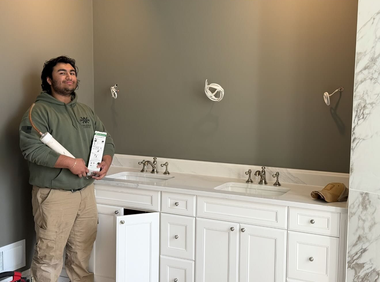 Man in a bathroom holding blueprints and a box; white vanity, gray wall, two sinks, marble trim.