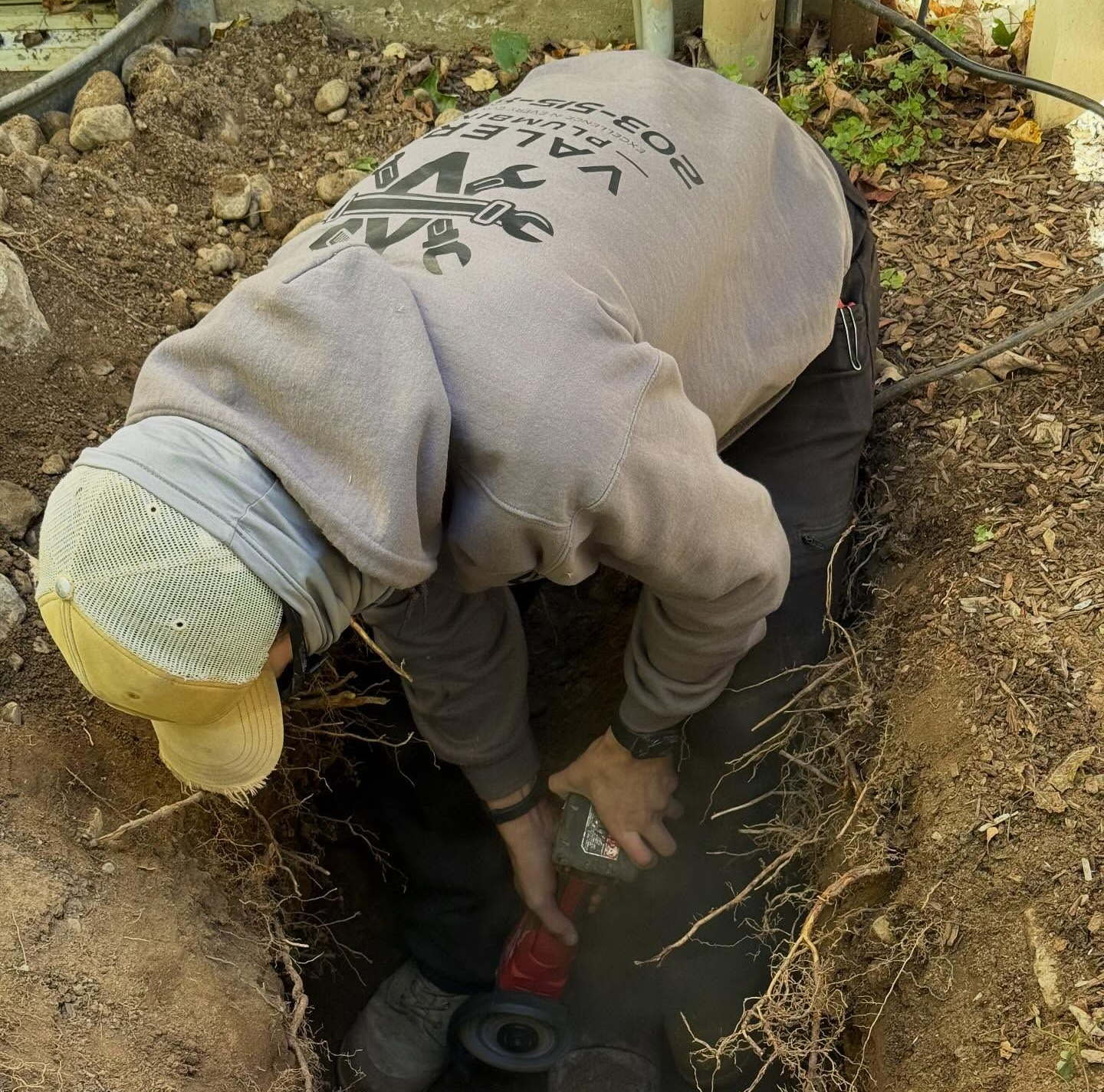 Person in a trench working on a pipe, wearing a light gray sweatshirt and a white cap.