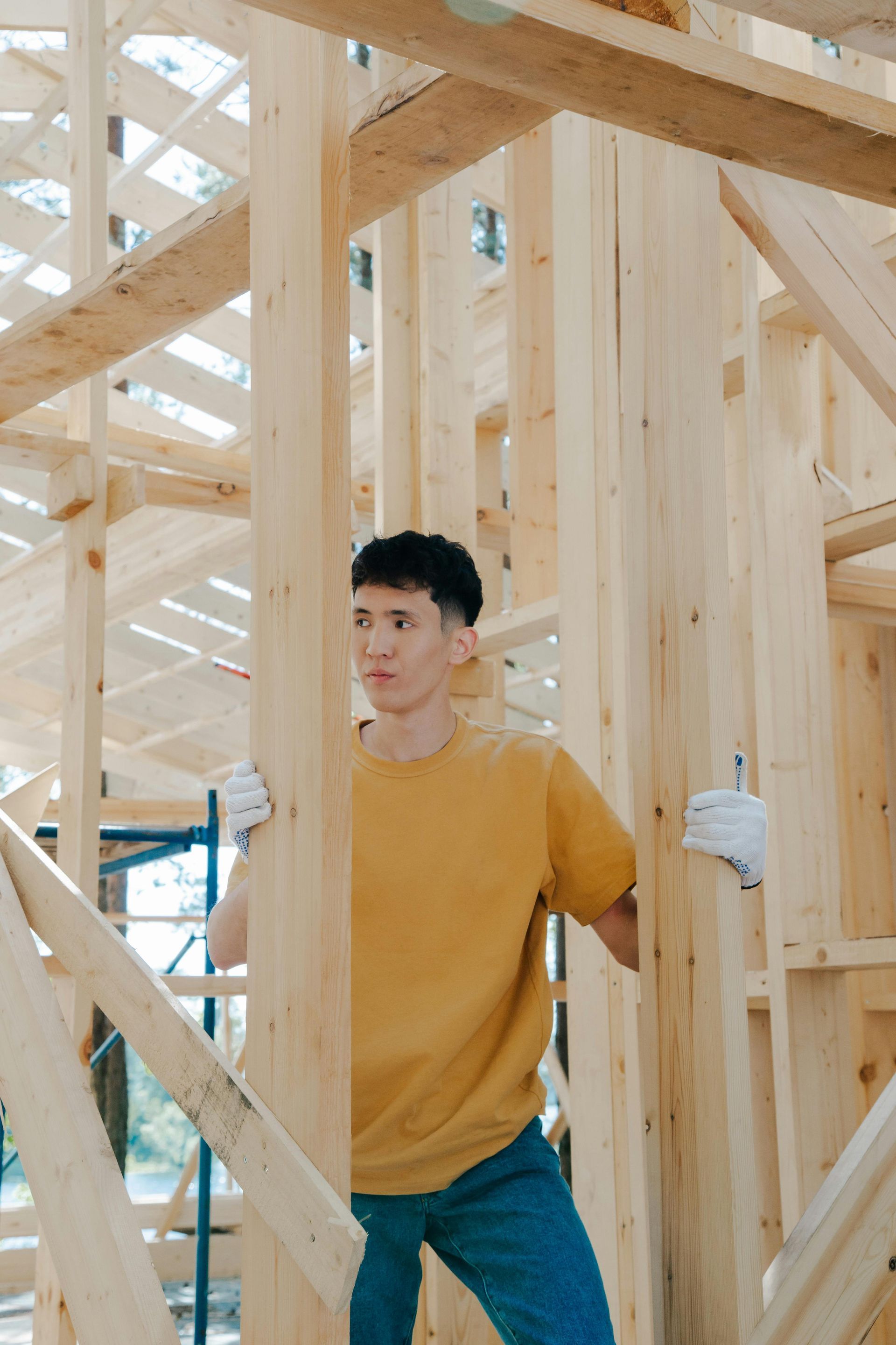 Man in yellow shirt and jeans inspecting wooden house framing, wearing gloves.