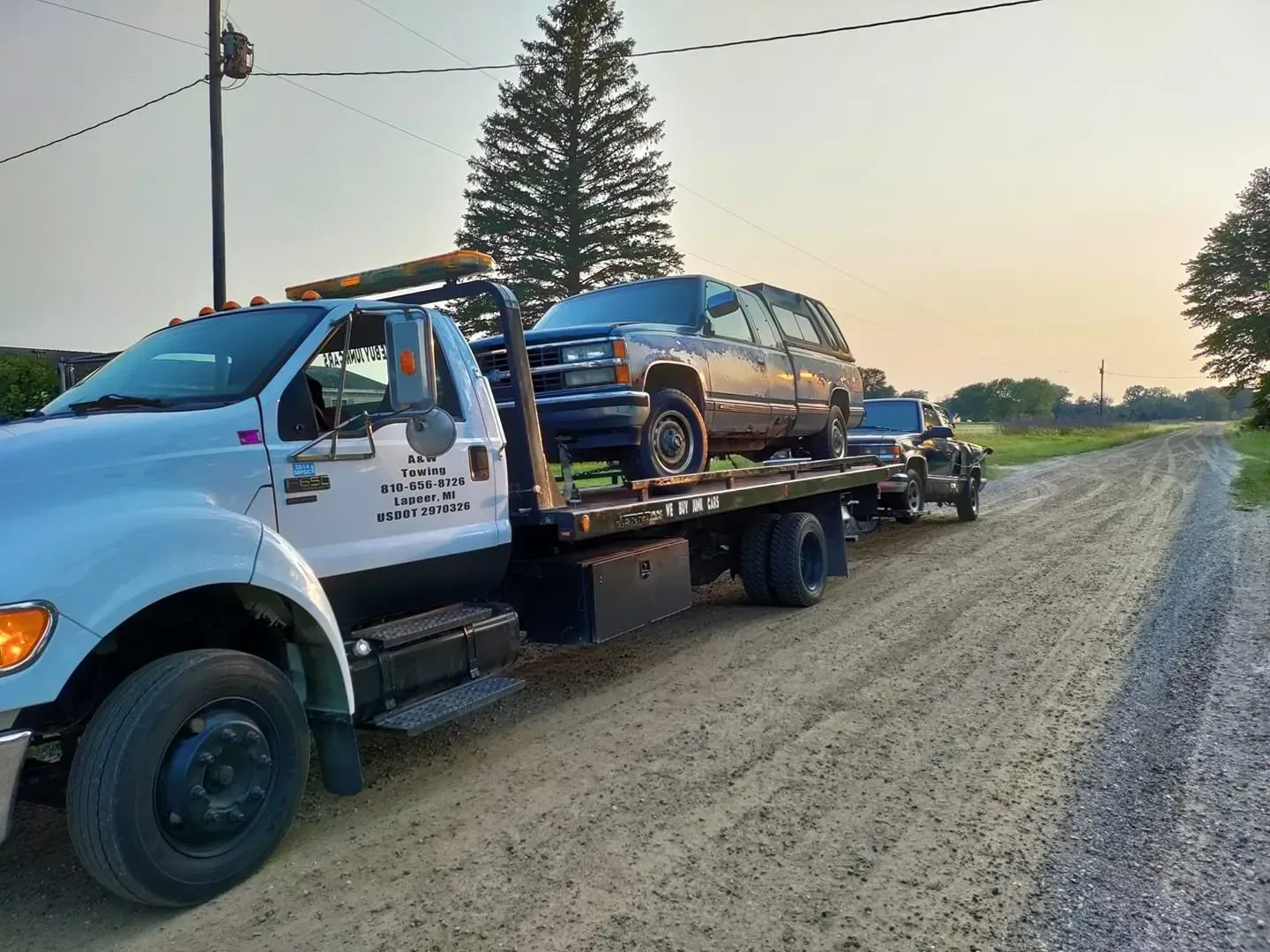 A tow truck is carrying two cars on a dirt road.