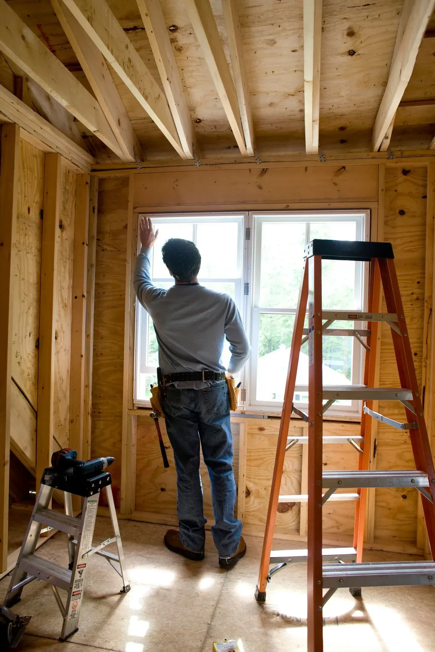 A man is standing in a room with a ladder and looking out a window.