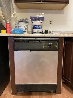 A stainless steel dishwasher under a countertop, with paint cans and supplies on top in a kitchen.