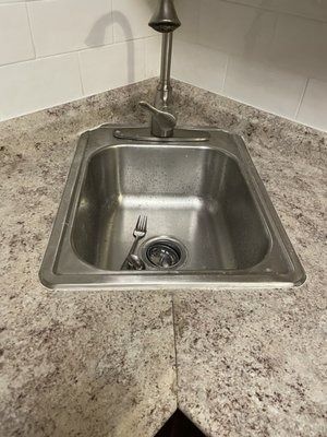 Stainless steel kitchen sink with faucet in a corner. A fork rests in the basin on a mottled gray countertop.