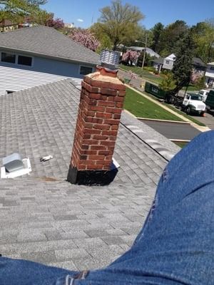 Brick chimney on a gray shingled roof, blue jeans in foreground, sunny day.
