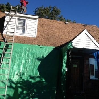 Roofer on roof, working. Green tarp covers house. Ladder leans against building.