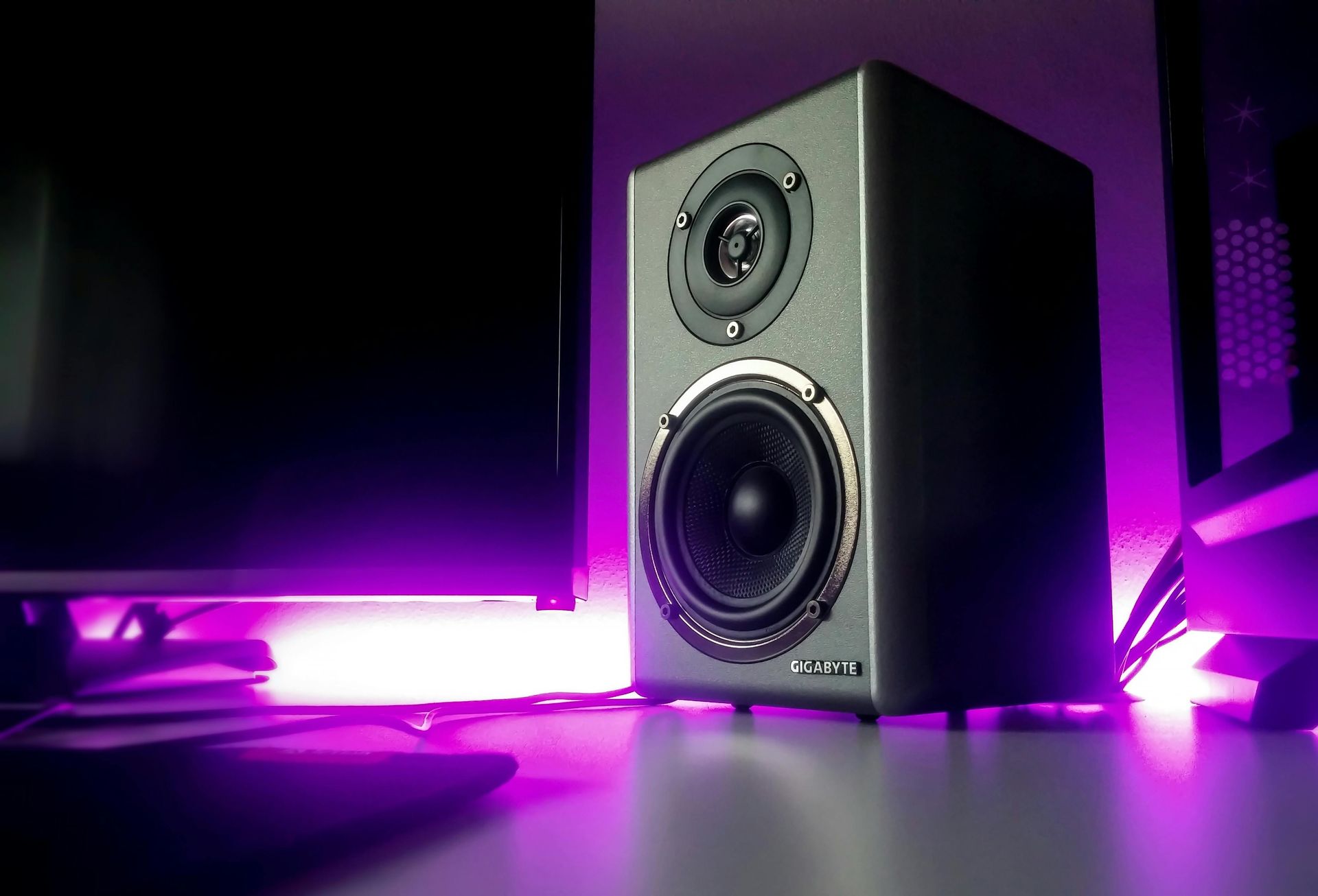 Altavoz negro sobre un escritorio, iluminado por luces violetas junto a un monitor.