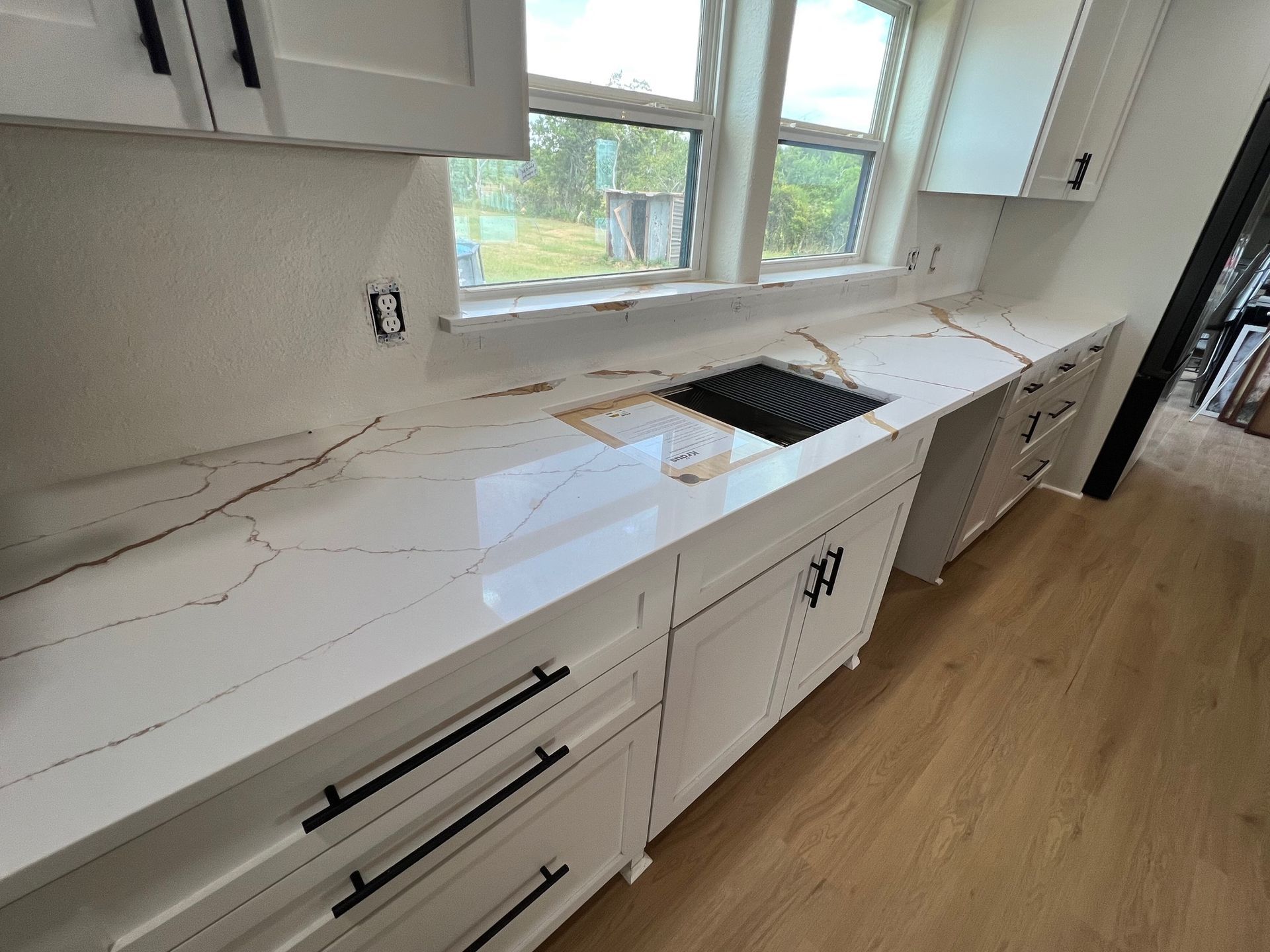 White kitchen cabinets and countertop with black hardware, sink, and window.