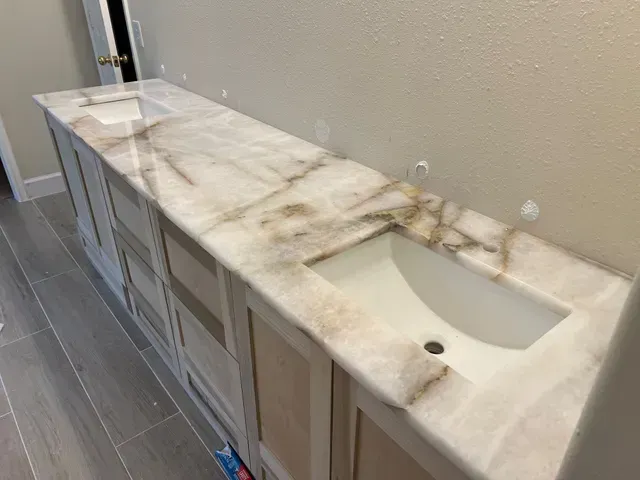 Long bathroom vanity with two sinks, marble countertop, light wood cabinets, beige walls.