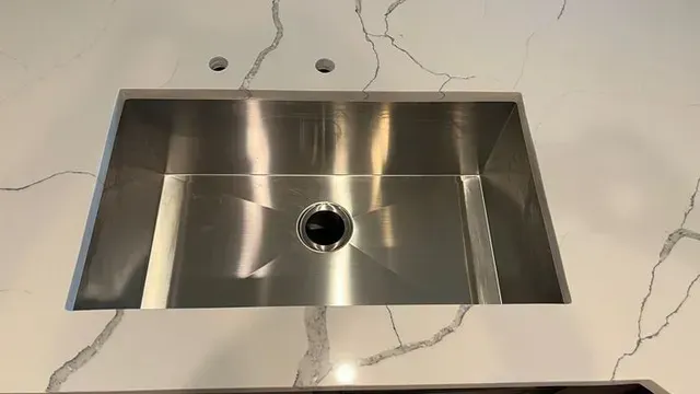 Stainless steel kitchen sink in a white countertop with a marble pattern. Two faucet holes above the sink.