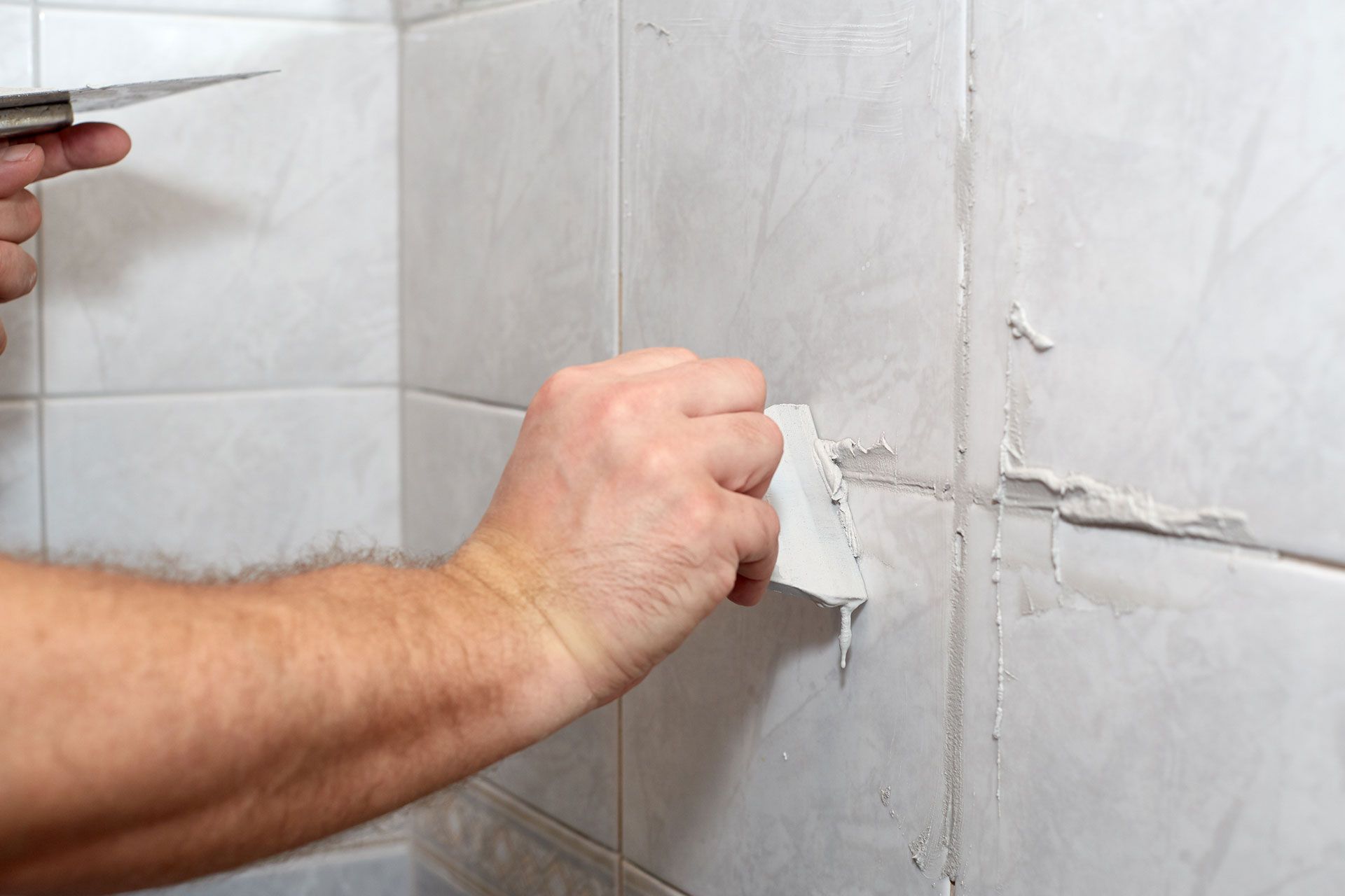 Person scraping peeling grout from white bathroom tiles with a putty knife.