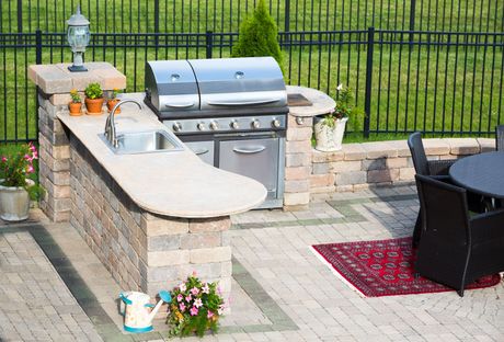 Outdoor stone patio kitchen with built-in stainless grill, sink, and curved countertop by a fence.