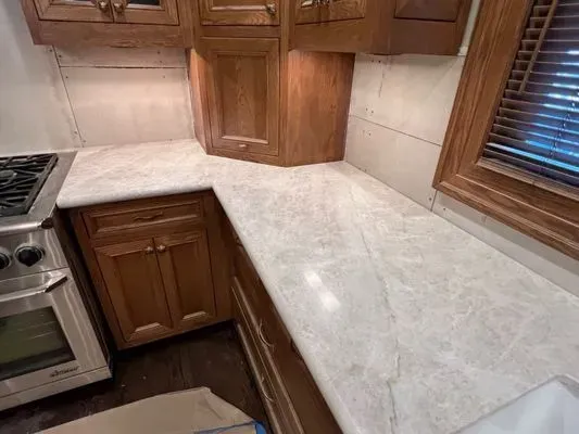 Kitchen countertop with marble-like pattern. Wooden cabinets below and above, stainless steel stove, and window with blinds.