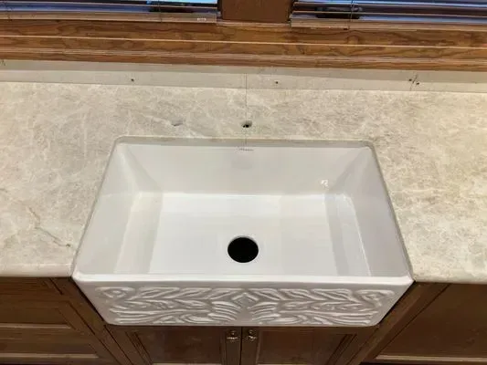White farmhouse sink in a cream-colored countertop with wood cabinets below, set beneath a wood-framed window.