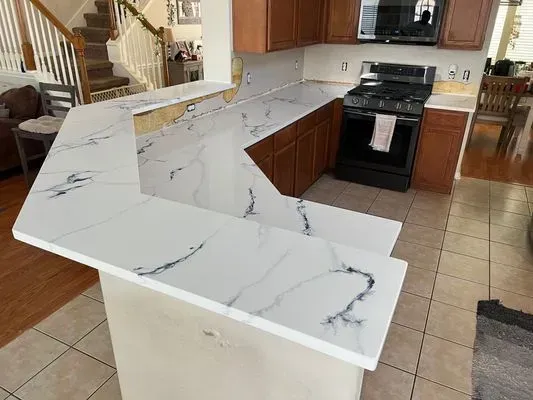 White marble-patterned kitchen countertops, dark cabinets, and gas stove in a kitchen with a view of a staircase.