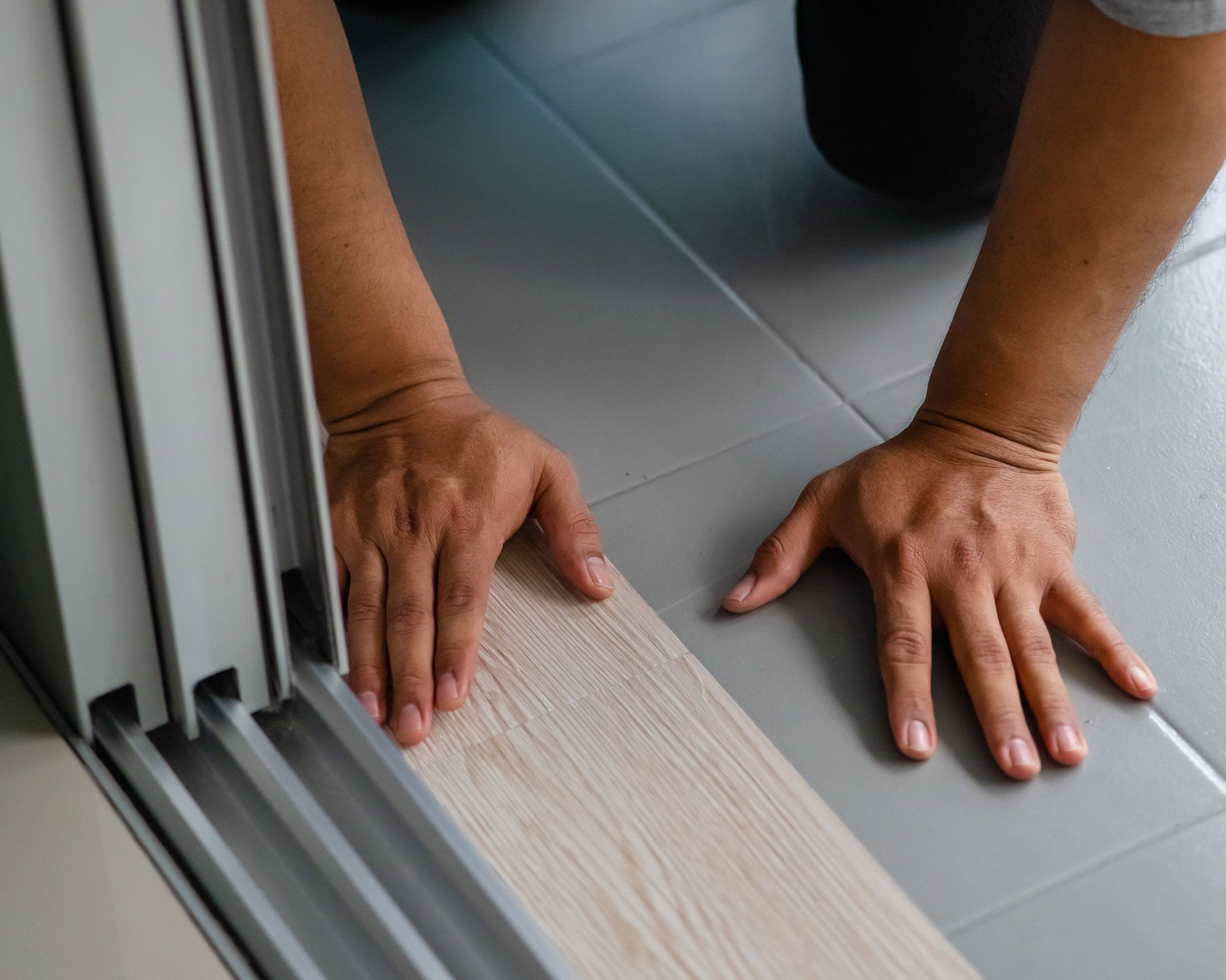 Hands installing wood-look flooring next to a door frame on a tile floor.
