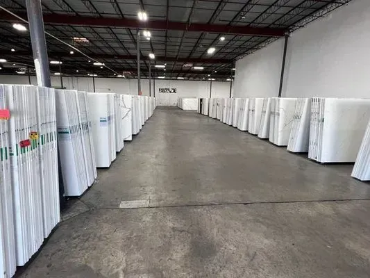 Warehouse interior with rows of large, white-wrapped slabs. Concrete floor, overhead lights, and a distant doorway.