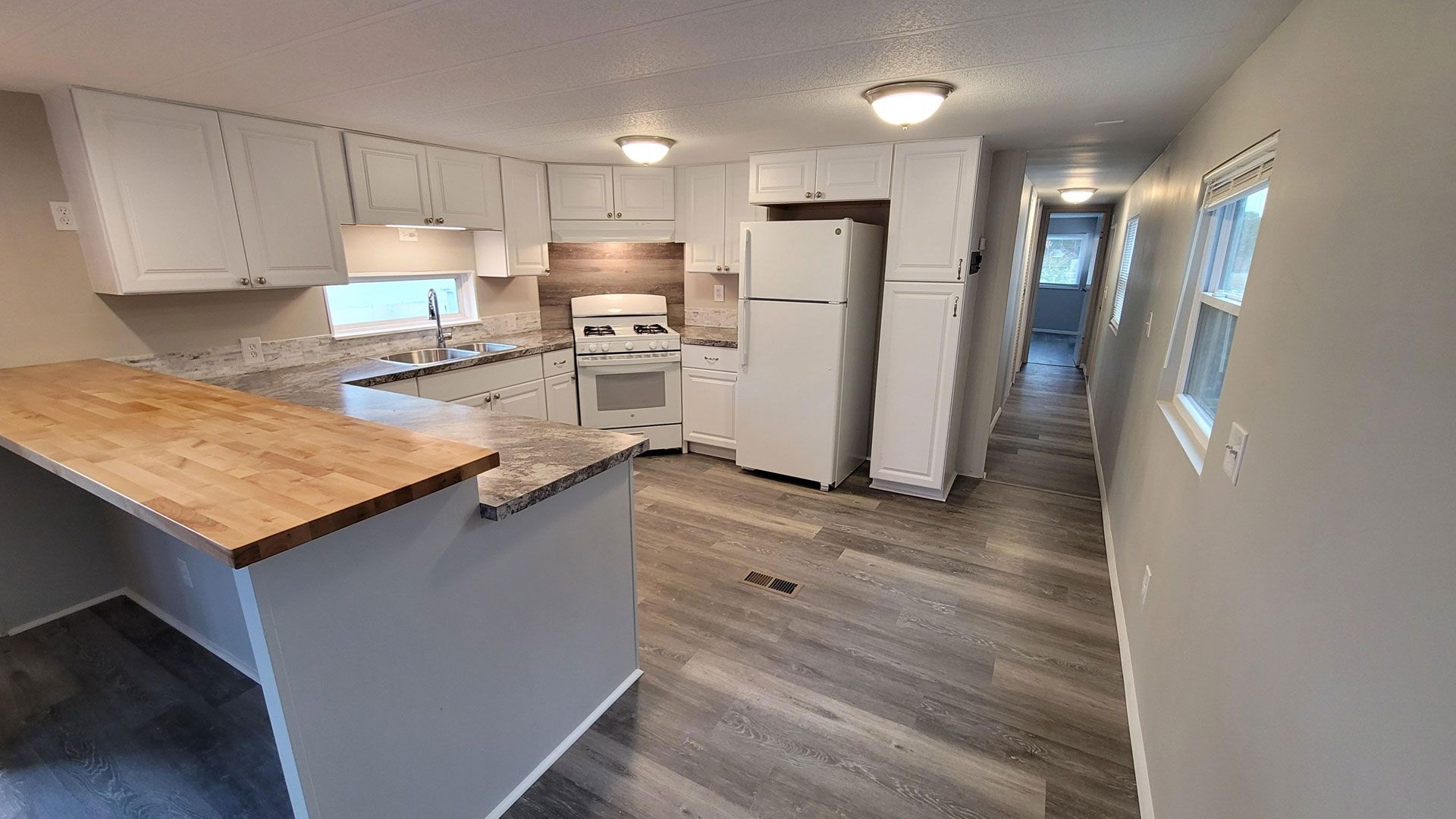 Empty white kitchen with wood countertops, gray flooring, white appliances, and windows on the right.