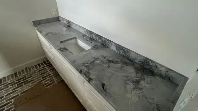 A gray marbled countertop installed on white cabinetry, next to a white wall and brick-patterned floor.