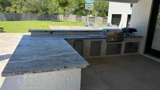 Outdoor kitchen with gray countertop, grill, and cabinets on a patio. Green yard in background.
