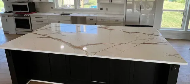 Kitchen island with white countertops and dark cabinets. Other white cabinets and windows in background.