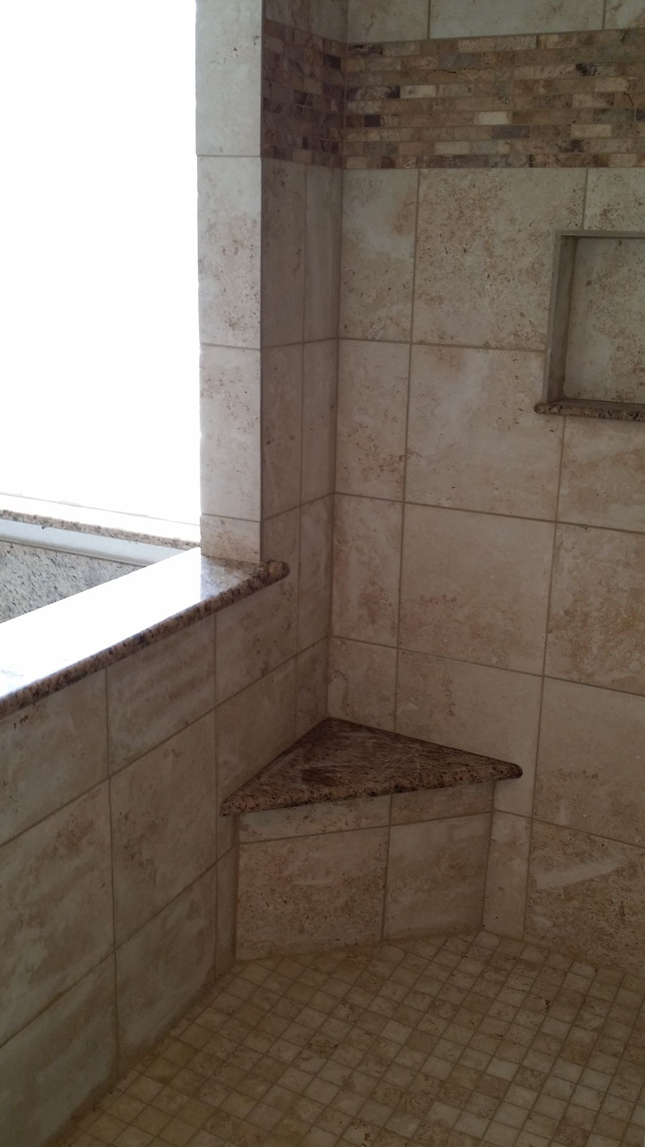 Corner of a shower with tiled walls and a built-in triangular bench. Neutral colors. Window visible.