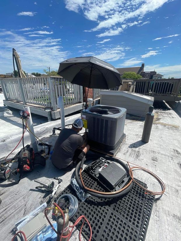 HVAC technician on a rooftop repairs an air conditioning unit, under an umbrella on a sunny day.