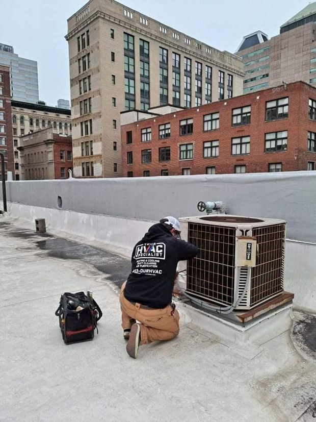 HVAC technician repairs an air conditioning unit on a city rooftop. Brown and grey buildings in the background.