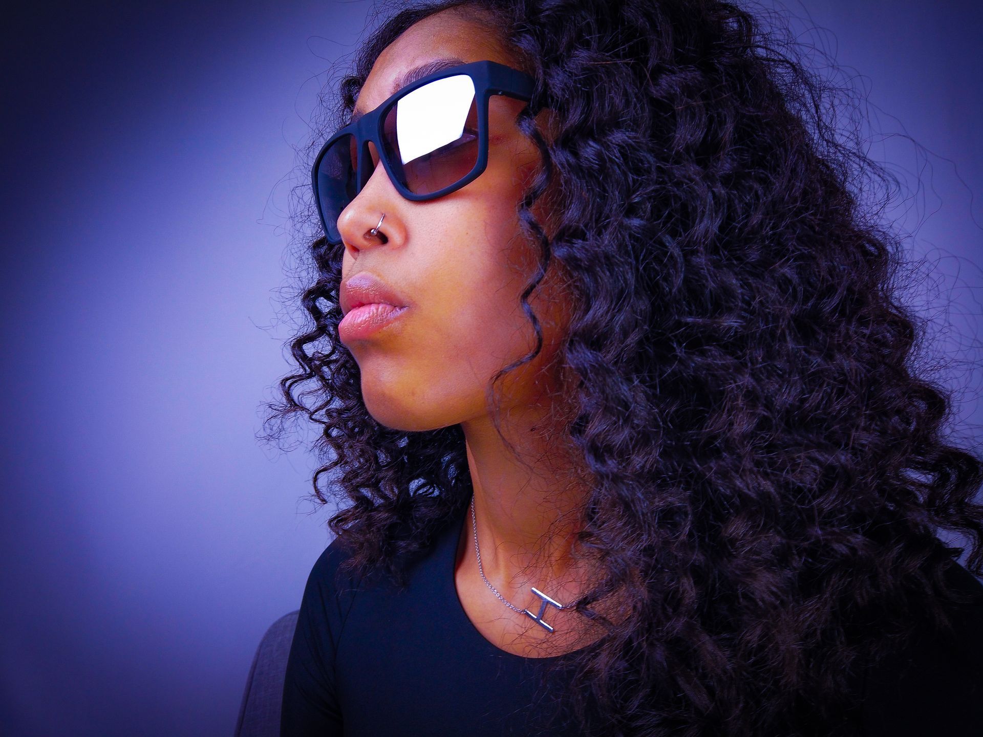 Person with curly hair wearing large sunglasses and a nose ring, looking to the side.
