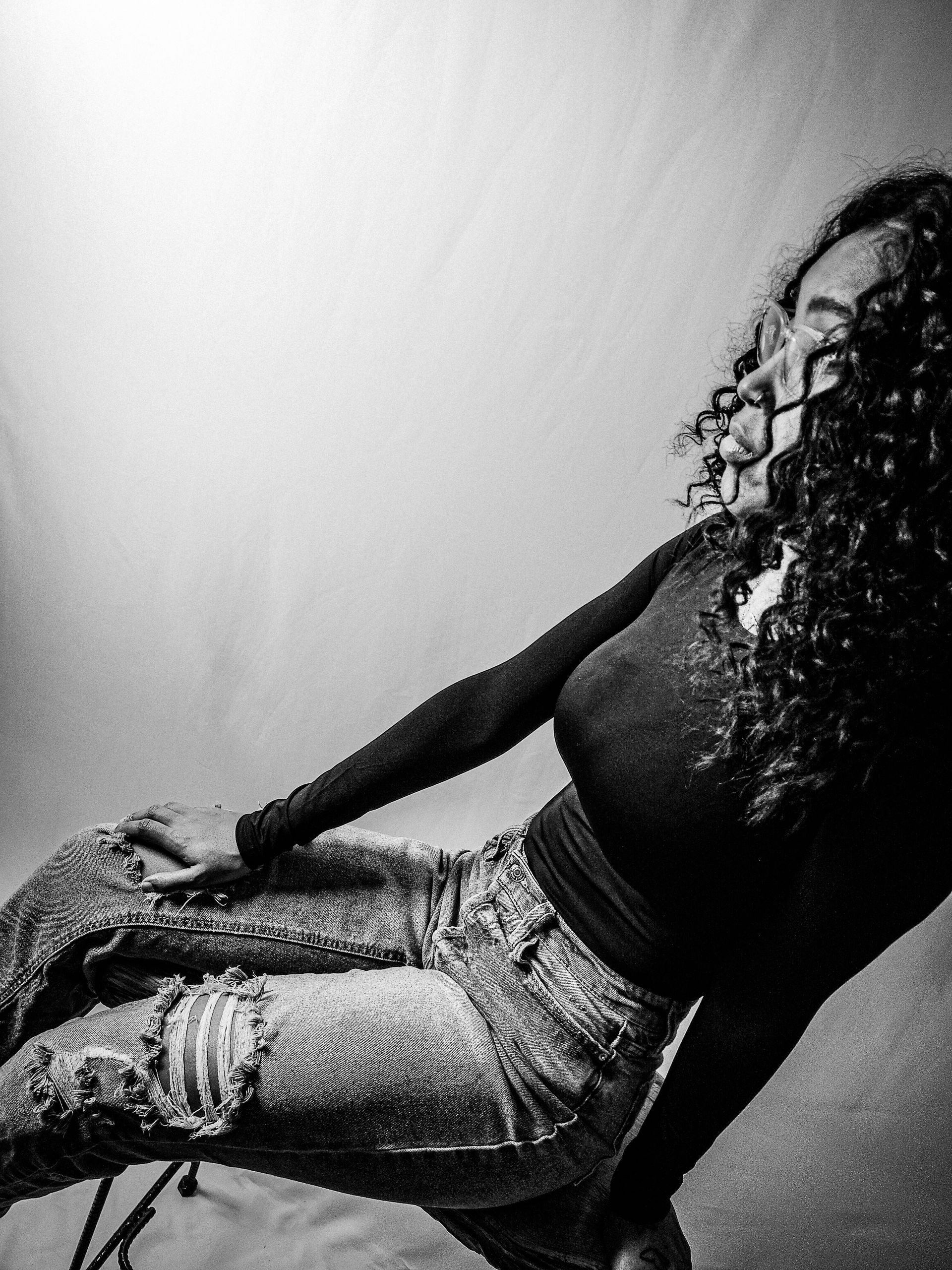 photo of person with curly hair sitting on a stool, wearing a long-sleeved top and ripped jeans, looking to the side.