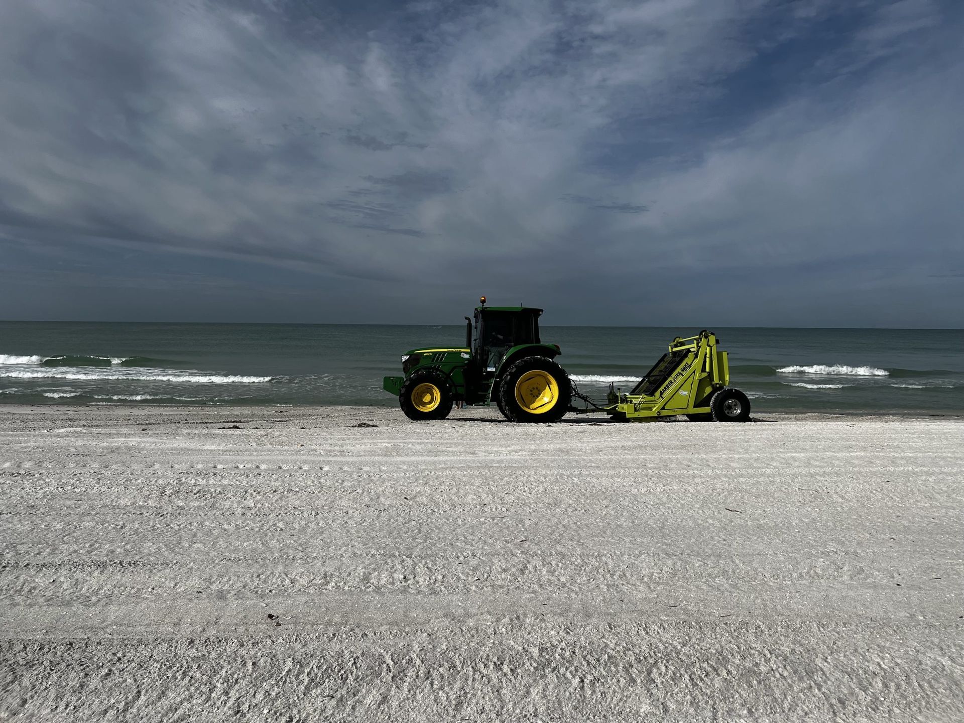 Green tractor with beach cleaner on a sandy beach near the ocean under a cloudy sky.