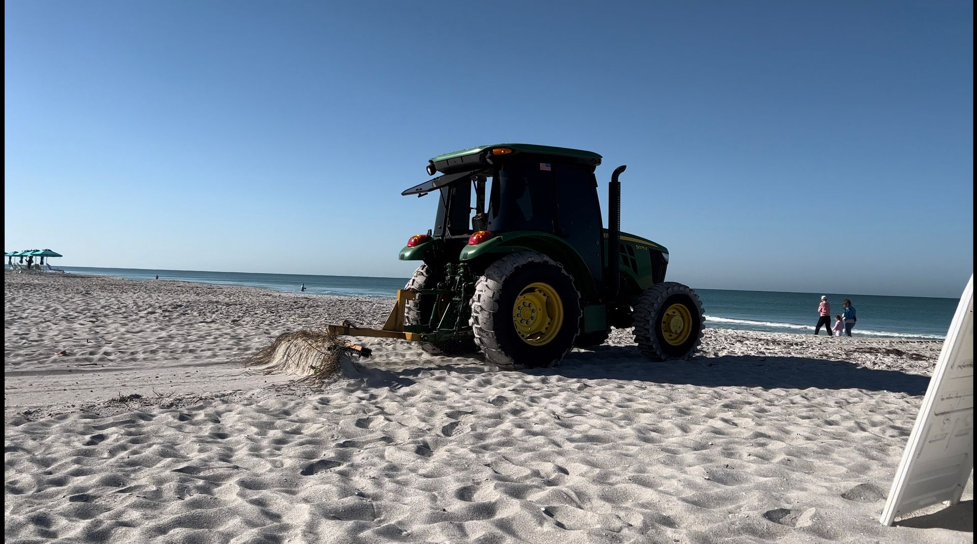 Green tractor raking sand on a beach with the ocean and a few people in the background.