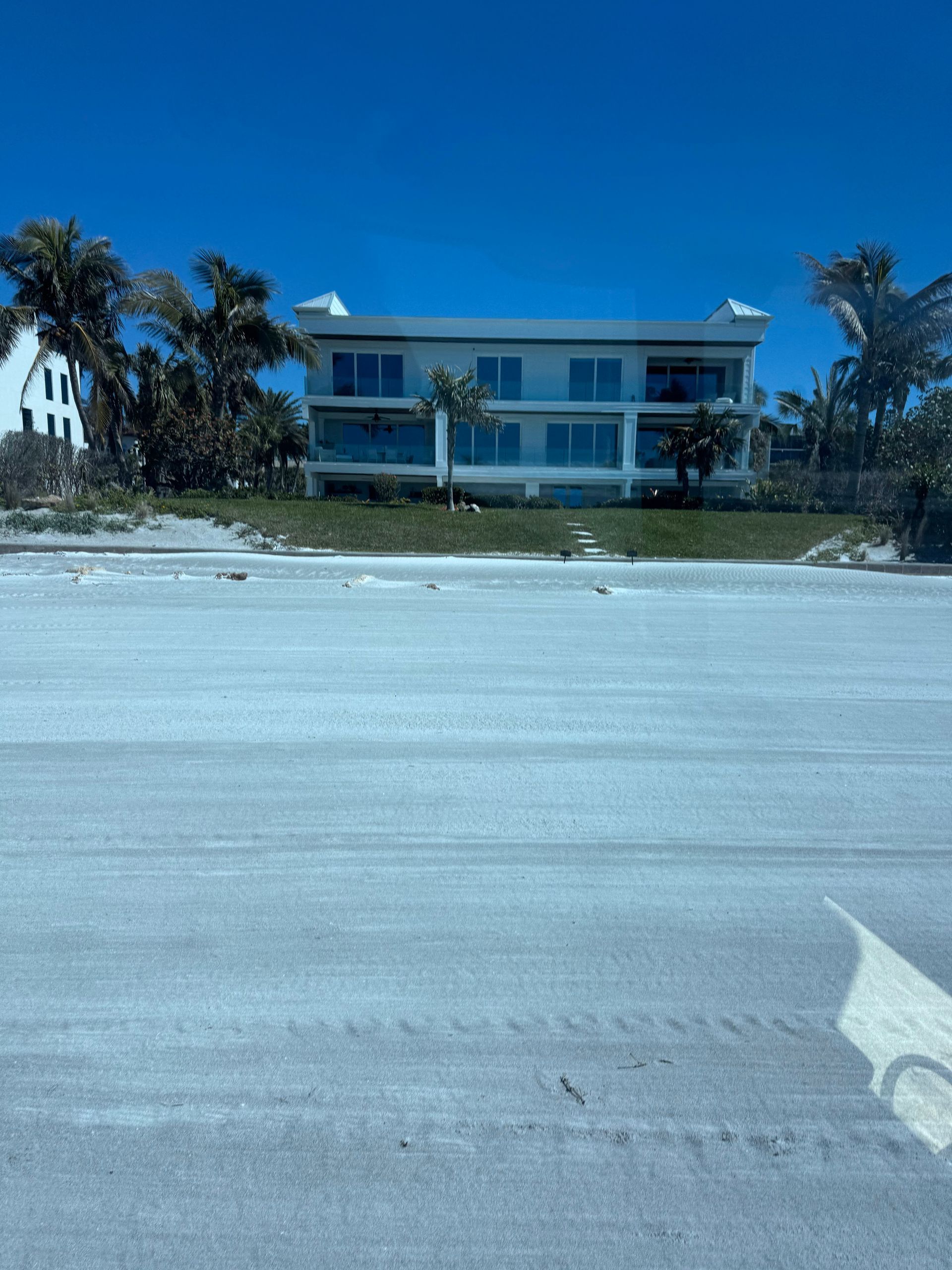 White beachfront house with glass balconies, on a sandy beach under a blue sky.