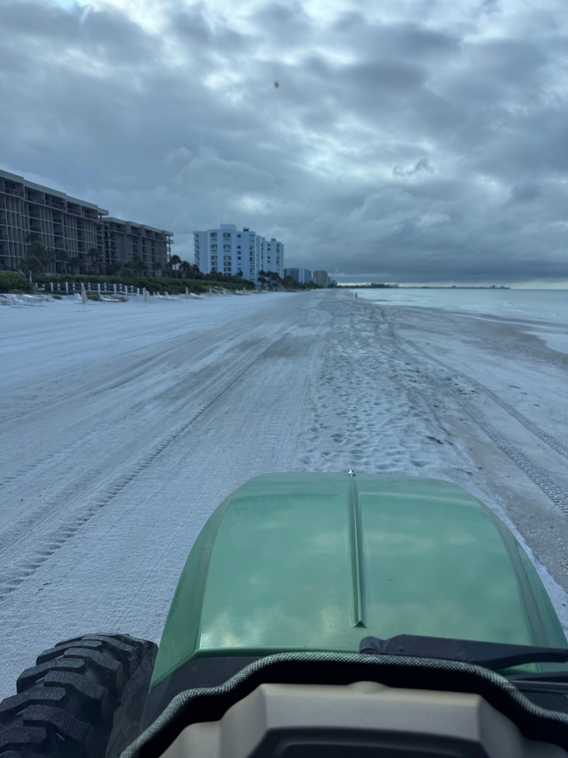 Green vehicle on a white sandy beach with buildings in the background under a cloudy sky.