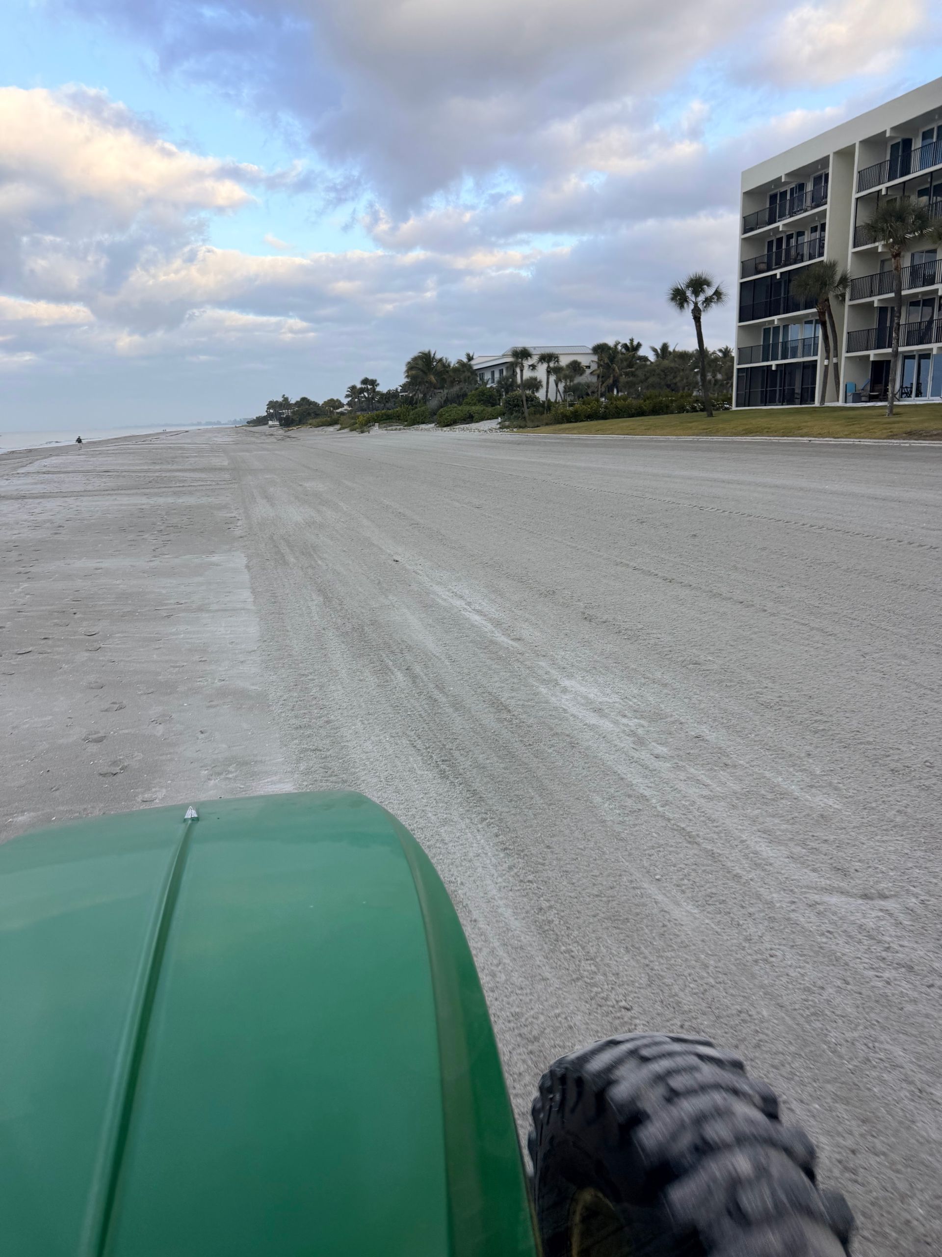 Green tractor driving on a sandy beach, buildings and cloudy sky in the background.