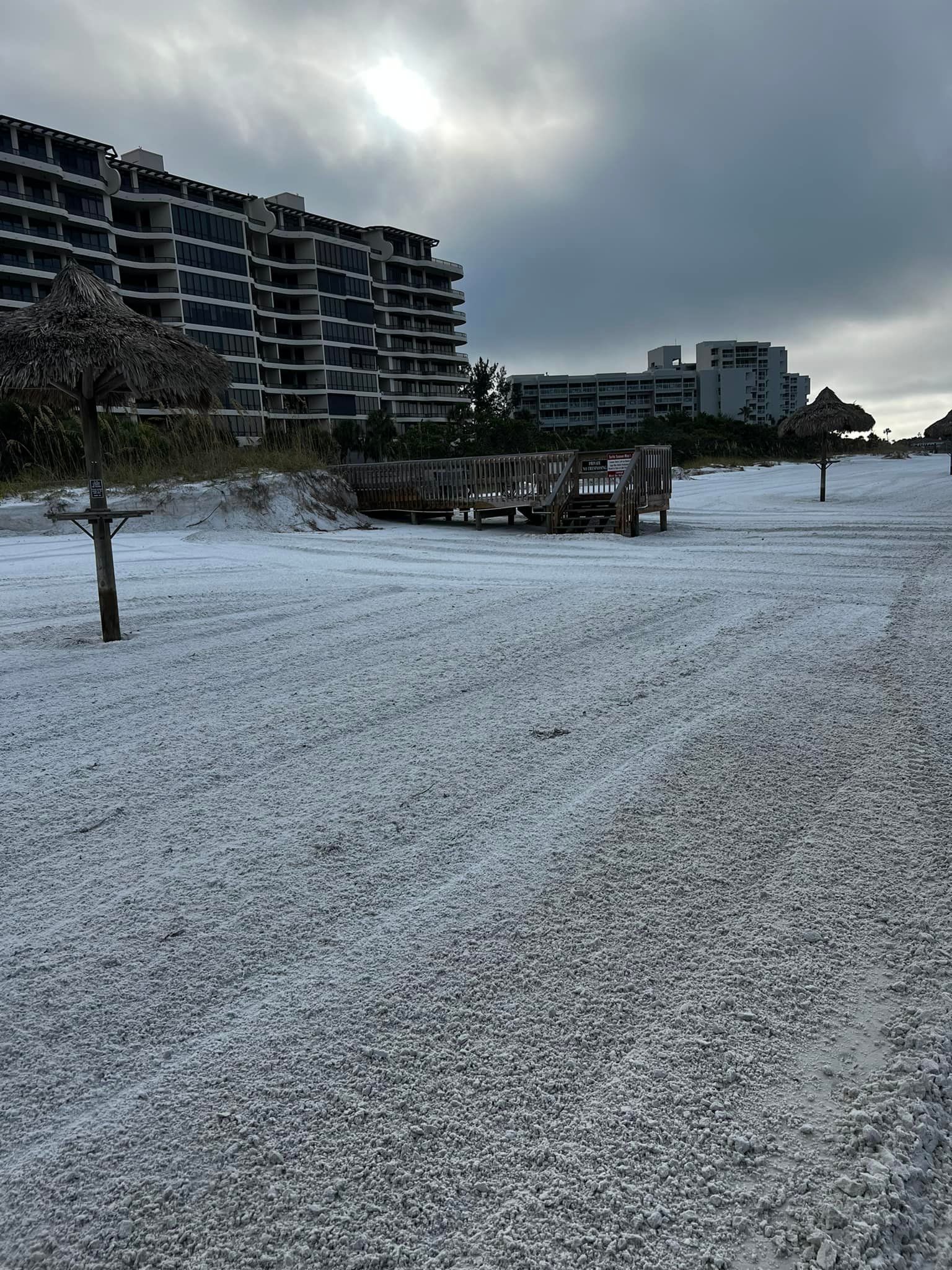 Beach covered in white debris, possibly hail. Buildings in the background under a cloudy sky.