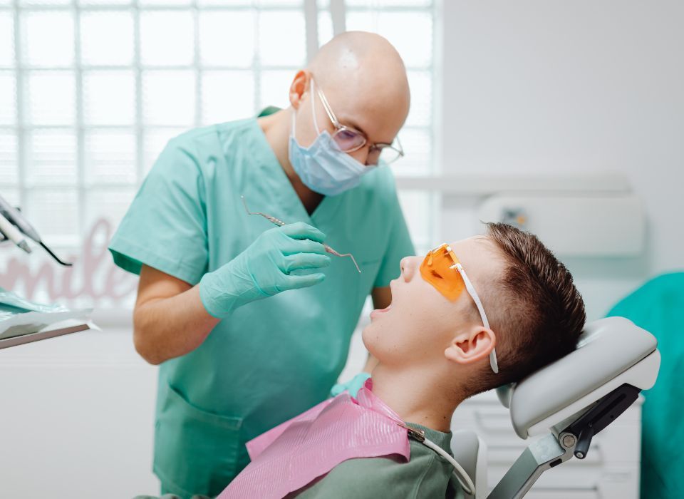 Dentist examining a patient's teeth in a dental chair. Patient wears orange safety glasses and bib.