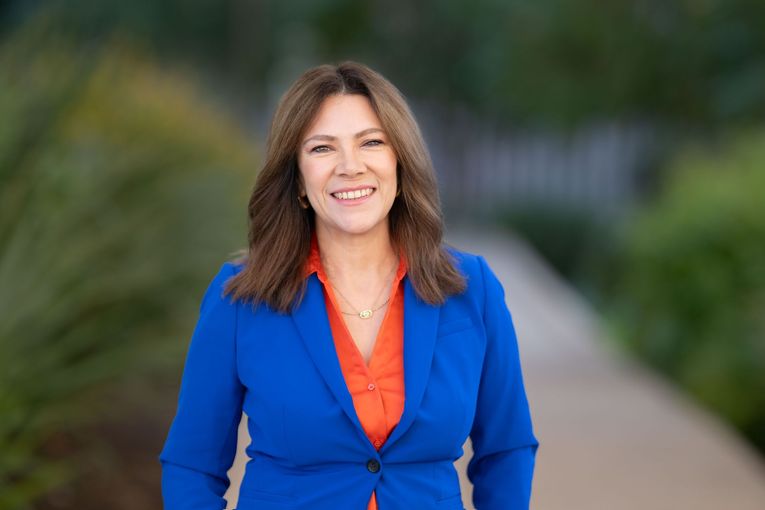 A woman wearing a blue and white jacket and orange earrings is smiling for the camera.