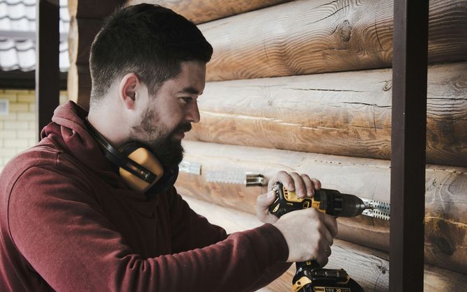 Man drilling into a log cabin wall with a power drill, wearing a maroon hoodie and headphones around his neck