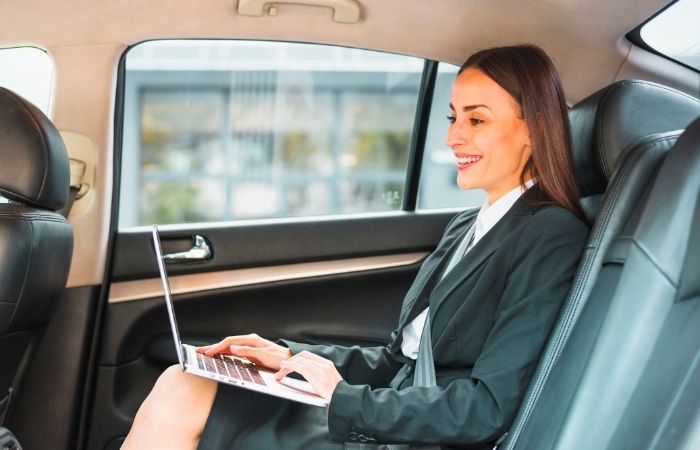 A woman in a suit is sitting in the back seat of a car using a laptop computer.