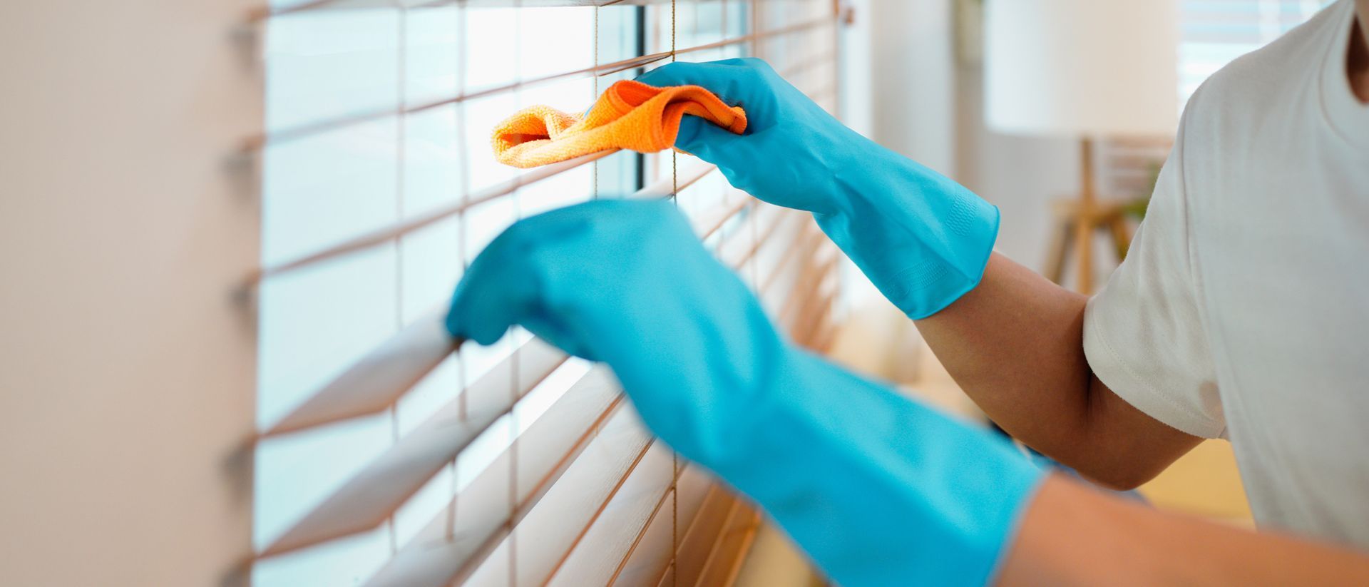 Blue-gloved hands cleaning window blinds with an orange cloth in a bright room Blue-gloved hands cleaning window blinds with an orange cloth in a bright room