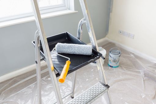 Paint supplies on a step ladder beside a window, with a roller tray and paint can on a drop cloth. Paint supplies on a step ladder beside a window, with a roller tray and paint can on a drop cloth.