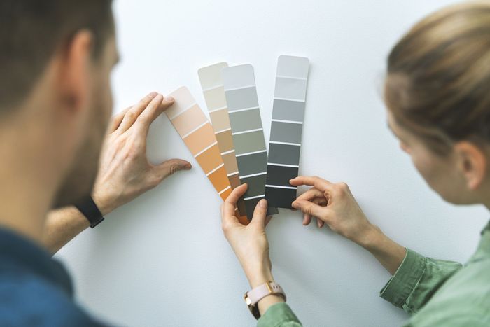 Two people compare paint swatches against a white wall, choosing neutral and warm color samples. Two people compare paint swatches against a white wall, choosing neutral and warm color samples.